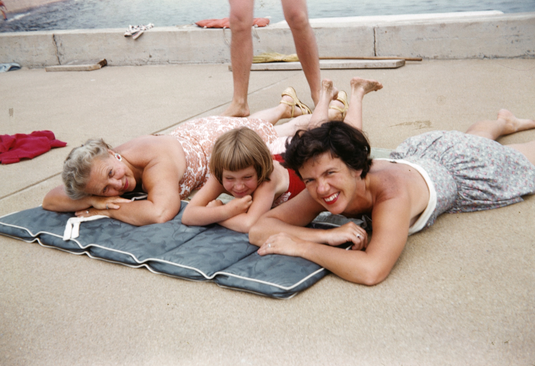 an old photo of two women and a child lying on a beach pad with water behind them