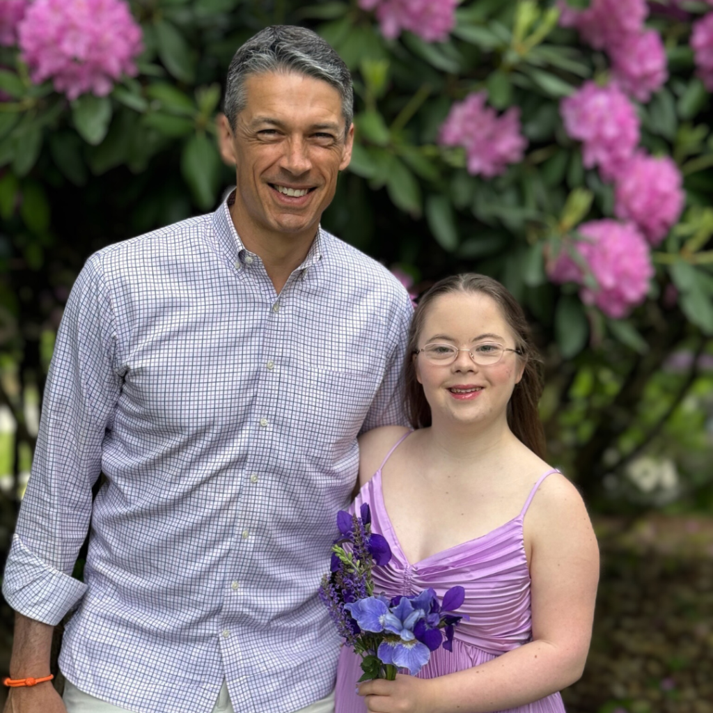 Peter and Penny smile for the camera in front of a flowering bush. Penny is wearing her purple prom dress and holding dark purple flowers.