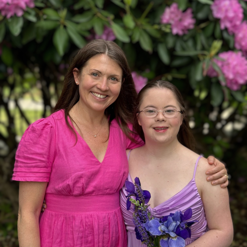Amy Julia and Penny smile for the camera in front of a flowering bush. Penny is wearing her purple prom dress and holding dark purple flowers.