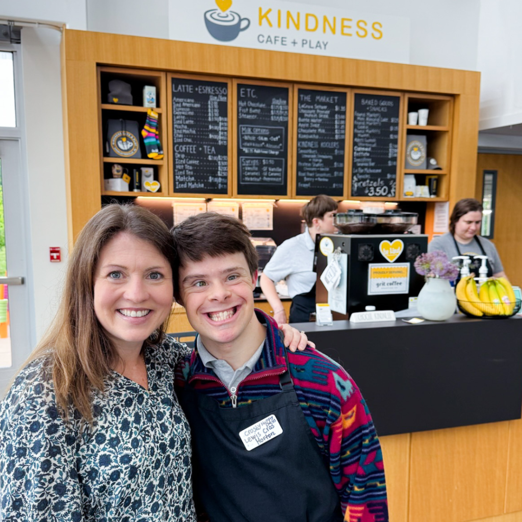 Amy Julia and a cafe employee stand in Kindness Cafe with their arms around each other and smile at the camera