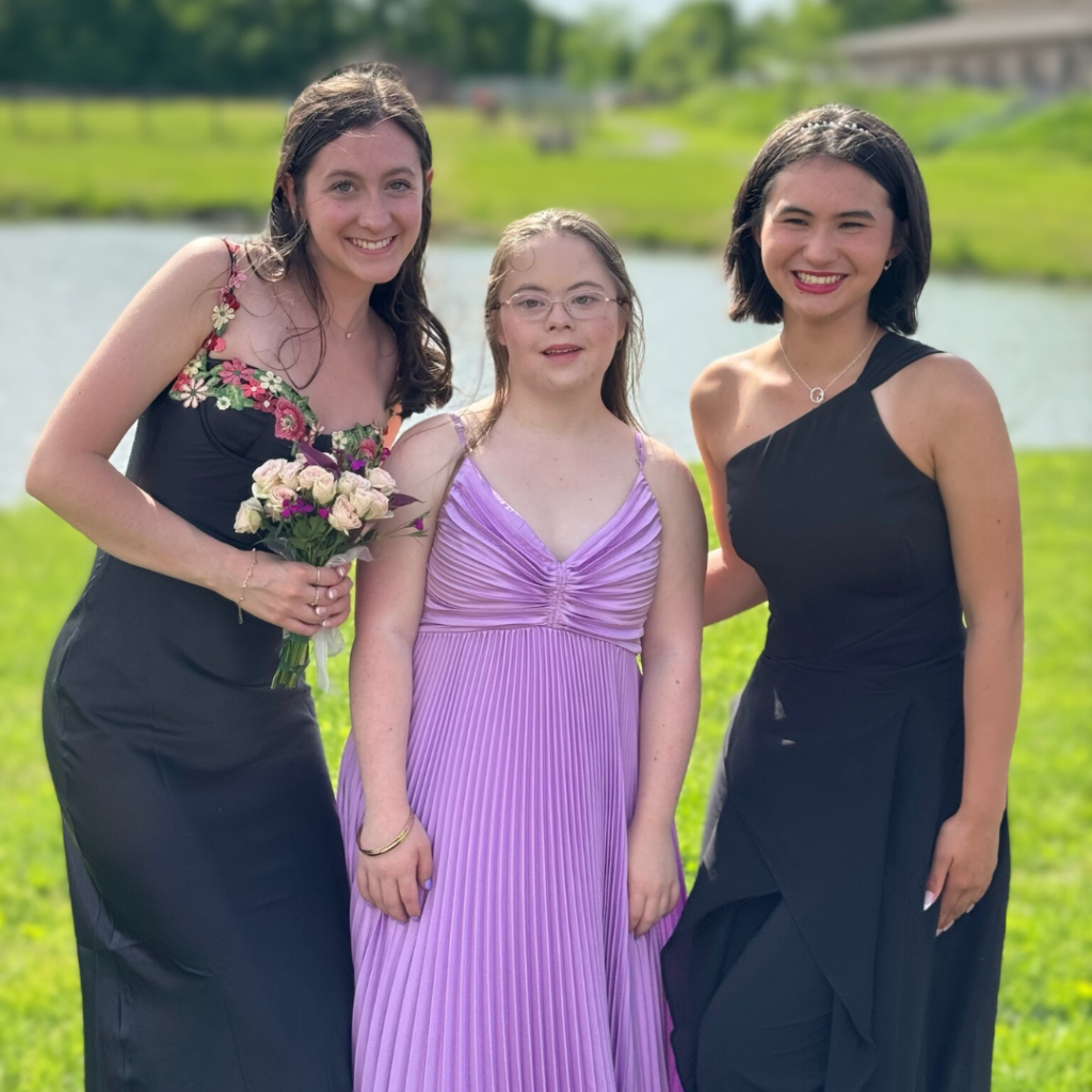 an outdoor photo of Penny wearing a purple prom dress and posing with two friends in front of a pond