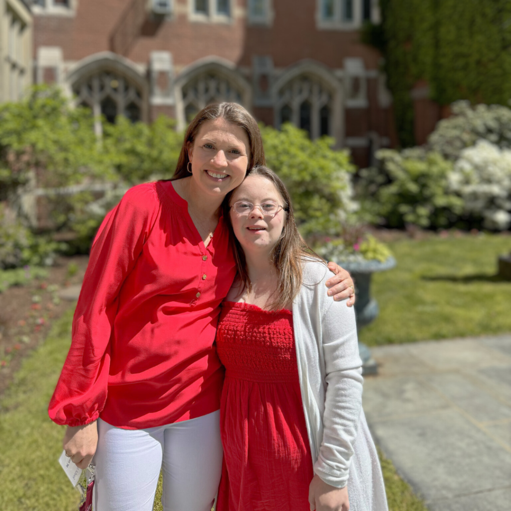 Amy Julia and Penny, both wearing red, stand in front of a garden and brick building