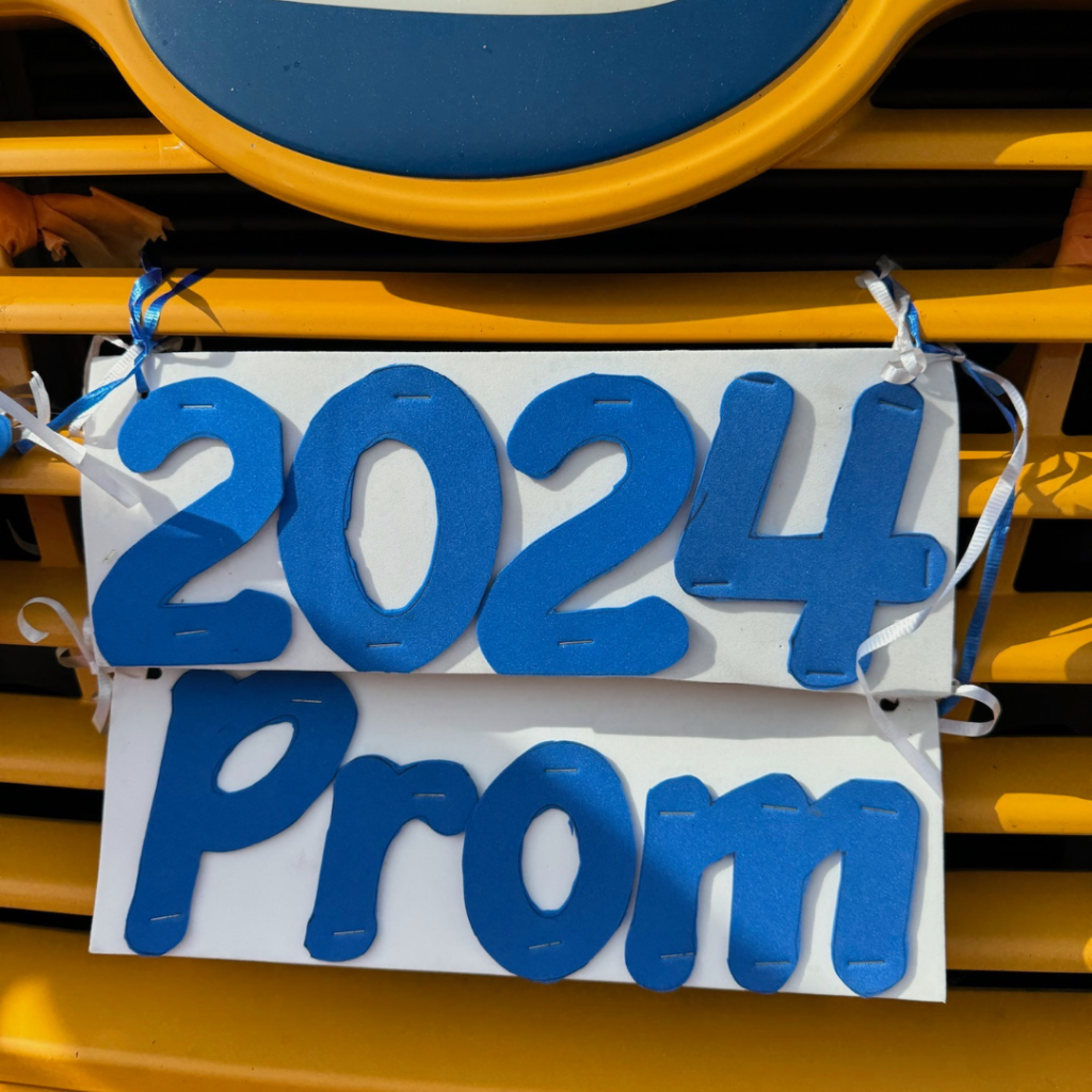 front grill of a yellow school bus with a white sign attached that says "2024 prom" in blue letters