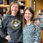 Katie Kishore and Amy Julia stand in Kindness Cafe with their arms around each other and smile at the camera