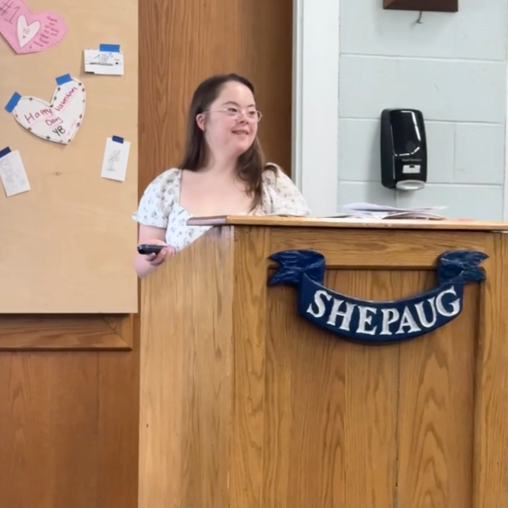 Penny smiles at her audience and stands at a podium in a classroom