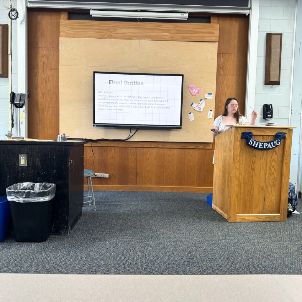 Penny stands at a classroom podium next to a TV with her presentation.