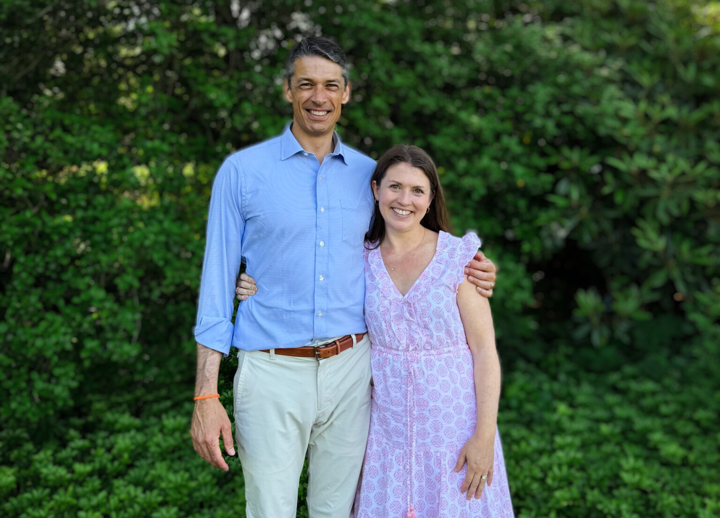 Peter and Amy Julia stand outside in front of green trees and bushes. They smile at the camera with their arms around each other