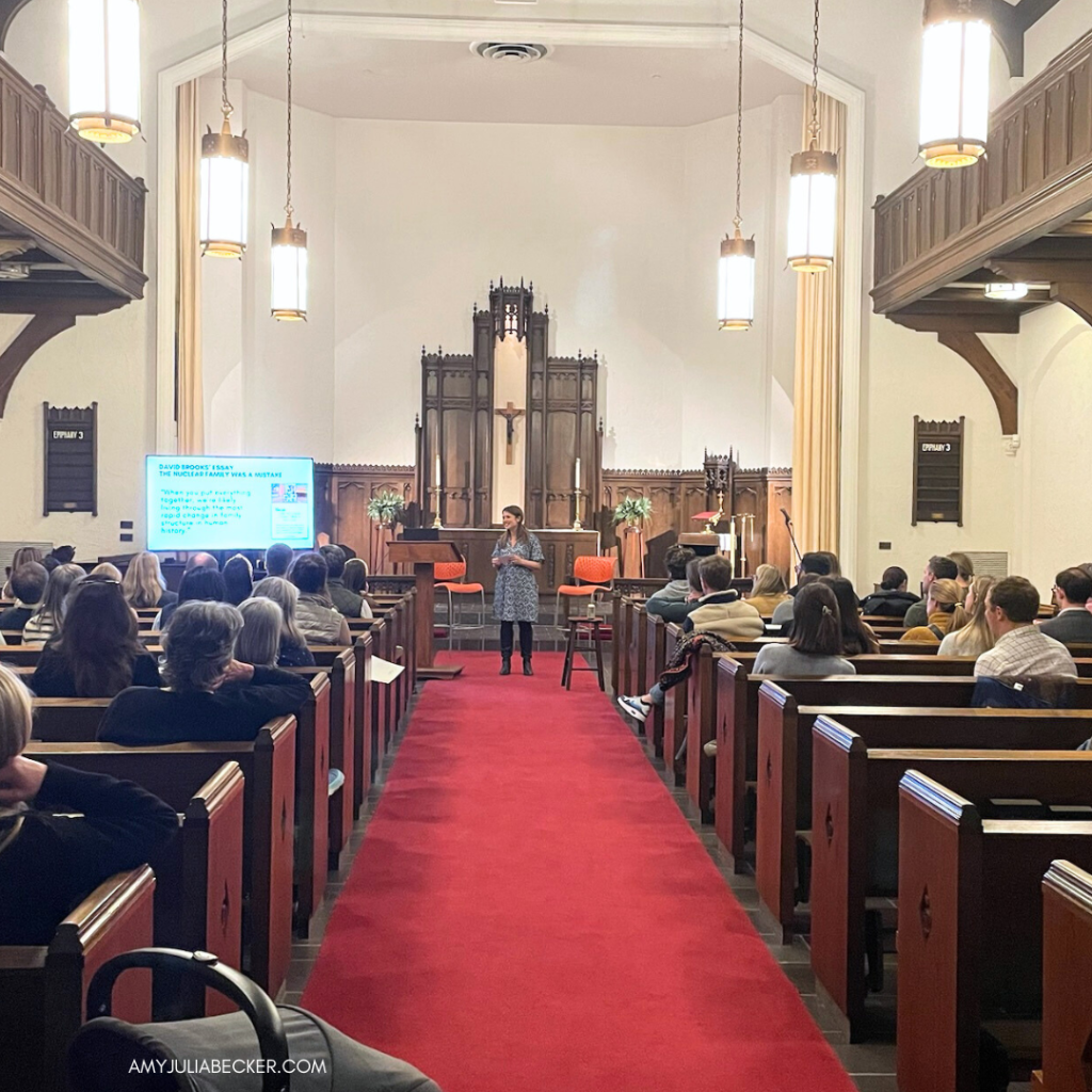 I’m speaking at the front of the church sanctuary, which has traditional architecture, featuring wooden pews, high ceilings with exposed beams, and large hanging lanterns.