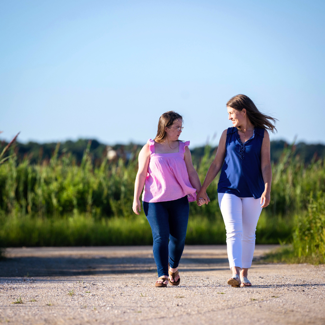 Penny and Amy Julia are walking hand in hand outdoors on a sunny day. Amy Julia is looking down at Penny and smiling warmly as they share a moment. They are walking along a dirt path with green grass and tall plants in the background, under a clear blue sky