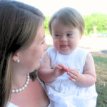 a photo in the middle is of Amy Julia smiles at baby Penny in her arms, who has Down syndrome and is wearing a white dress and looking back with a playful expression. They are outside on a sunny day, with greenery in the background. On either side of the photo are screenshots of a new study about developmental milestones for children with Down syndrome