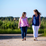 Penny and Amy Julia are walking hand in hand outdoors on a sunny day. Amy Julia is looking down at Penny and smiling warmly as they share a moment. They are walking along a dirt path with green grass and tall plants in the background, under a clear blue sky