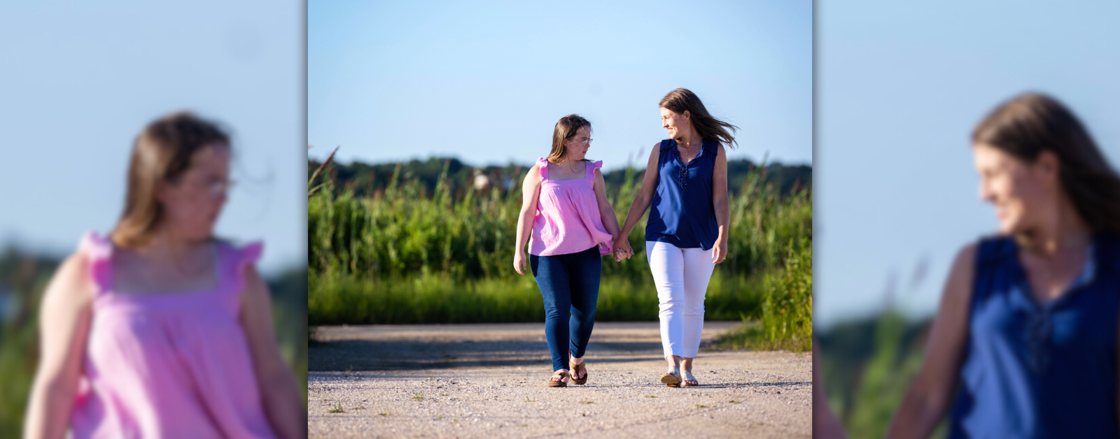 Penny and Amy Julia are walking hand in hand outdoors on a sunny day. Amy Julia is looking down at Penny and smiling warmly as they share a moment. They are walking along a dirt path with green grass and tall plants in the background, under a clear blue sky