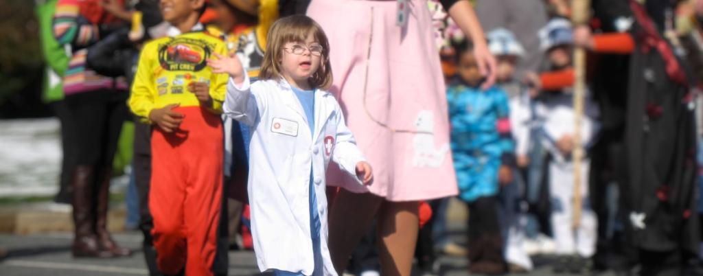 A photo from 2011 of Penny, who has Down syndrome, dressed as a doctor in a white lab coat, walking and waving during a parade. She is participating with other children, who are dressed in various colorful costumes