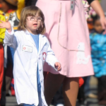 A photo from 2011 of Penny, who has Down syndrome, dressed as a doctor in a white lab coat, walking and waving during a parade. She is participating with other children, who are dressed in various colorful costumes