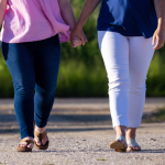 Penny and Amy Julia walking side by side on a dirt path, holding hands. One is wearing a pink top with dark blue jeans, and the other is wearing a blue top with white pants. Both are wearing sandals, and the image captures them mid-step.