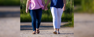 Penny and Amy Julia walking side by side on a dirt path, holding hands. One is wearing a pink top with dark blue jeans, and the other is wearing a blue top with white pants. Both are wearing sandals, and the image captures them mid-step.