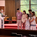 Amy Julia's family stands at the front of the church facing the congregation. The pastor stands next to them reading