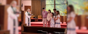 Amy Julia's family stands at the front of the church facing the congregation. The pastor stands next to them reading