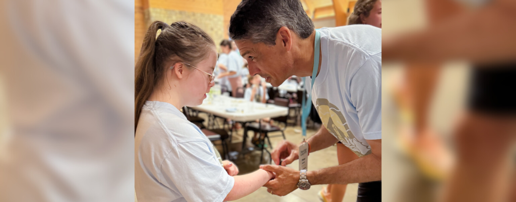 s the shape of a cross on her hands. He is leaning in slightly. Both are wearing light blue Hope Heals T-shirts. In the background, other people can be seen in a room with tables, creating a warm, communal atmosphere. The mood of the scene is caring and supportive.