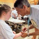 s the shape of a cross on her hands. He is leaning in slightly. Both are wearing light blue Hope Heals T-shirts. In the background, other people can be seen in a room with tables, creating a warm, communal atmosphere. The mood of the scene is caring and supportive.