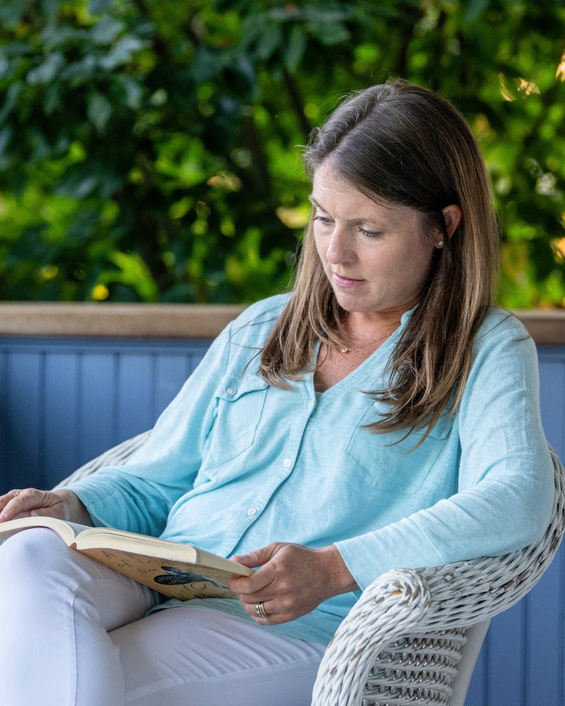 Amy Julia sits in a wicker chair on a porch reading a book