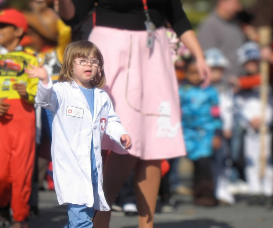 A photo from 2011 of Penny, who has Down syndrome, dressed as a doctor in a white lab coat, walking and waving during a parade. She is participating with other children, who are dressed in various colorful costumes