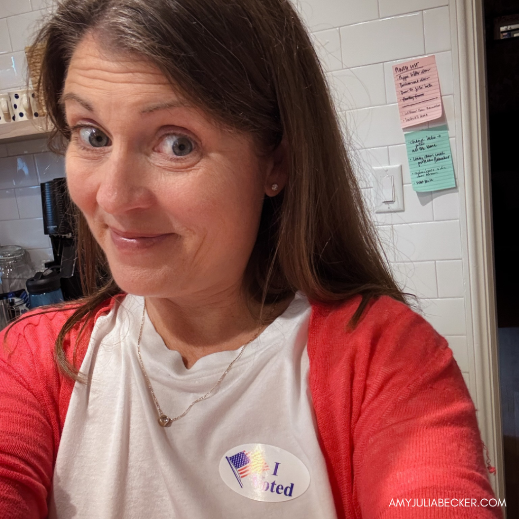 Amy Julia smiles for a selfie. An "I Voted" sticker is on her shirt. The background features a wall with colorful sticky notes.