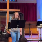 Penny stands at the front of the church holding a mic and leading the congregation in reading the Lord's prayer