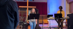 Penny stands at the front of the church holding a mic and leading the congregation in reading the Lord's prayer