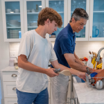 Amy Julia's family working together in a kitchen: Peter and William unload the dishwasher, while Amy Julia and Penny are in the background.