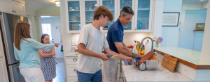 Amy Julia's family working together in a kitchen: Peter and William unload the dishwasher, while Amy Julia and Penny are in the background.