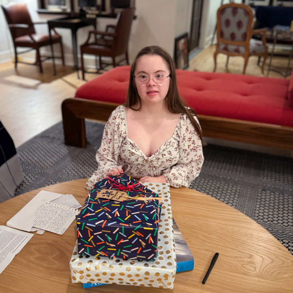 Penny sits at a wooden table with neatly wrapped presents in colorful wrapping paper in front of her.