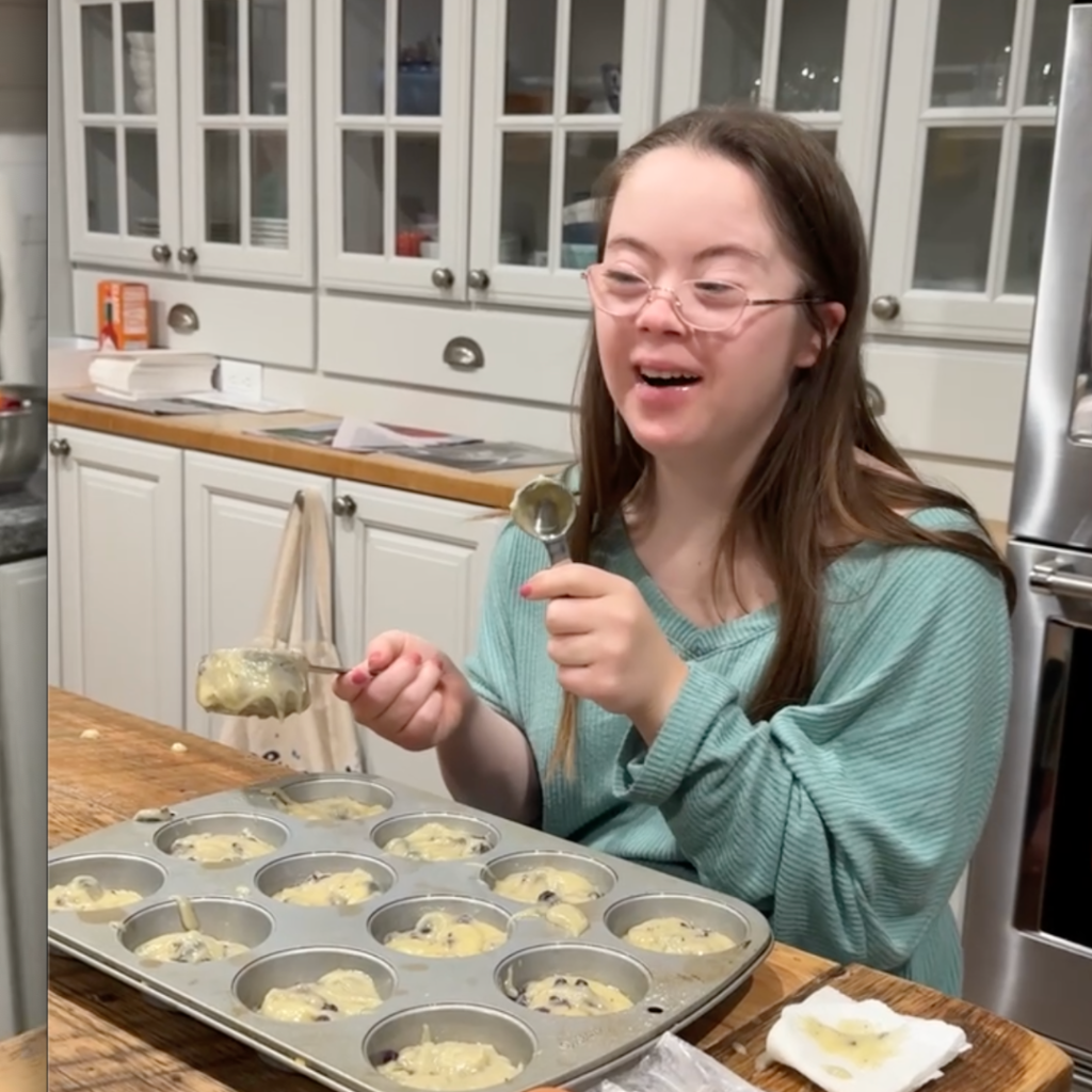 Penny sitting at a kitchen counter smiling widely with a filled muffin tin in front of her