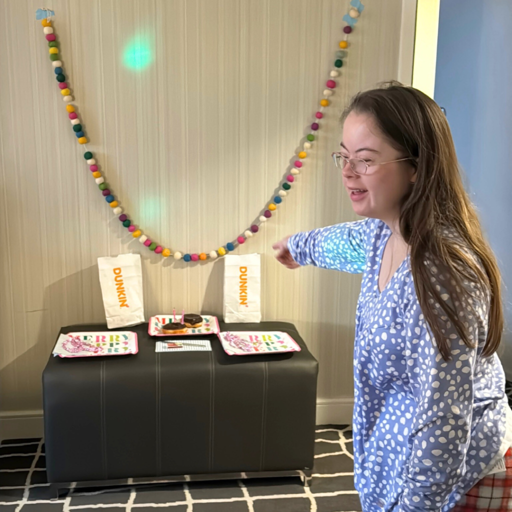 A festive setup with colorful string decorations on the wall. Penny in blue polka-dotted pajamas points toward a table adorned with party plates, and Dunkin Donuts bags.