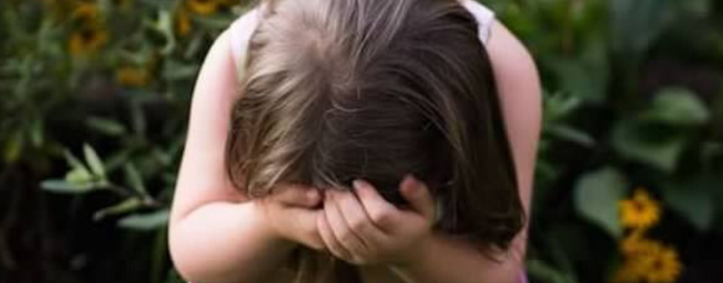 a 2012 photo of Penny crouching in the grass in a garden, covering her face with both hands. Behind her, there is a backdrop of yellow flowers and dark green foliage