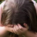 a 2012 photo of Penny crouching in the grass in a garden, covering her face with both hands. Behind her, there is a backdrop of yellow flowers and dark green foliage