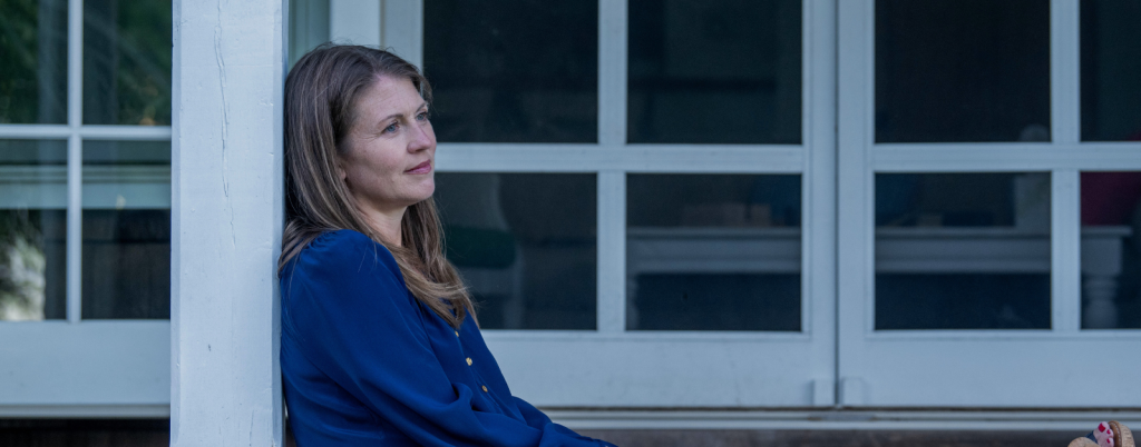 Amy Julia sits against a white pillar on a porch, gazing into the distance with a contemplative expression. The background features glass doors with a dark interior visible through them.