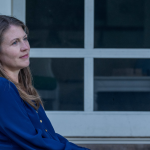 Amy Julia sits against a white pillar on a porch, gazing into the distance with a contemplative expression. The background features glass doors with a dark interior visible through them.