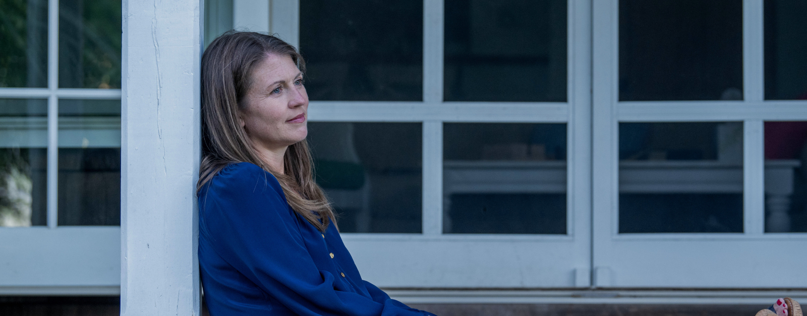 Amy Julia sits against a white pillar on a porch, gazing into the distance with a contemplative expression. The background features glass doors with a dark interior visible through them.