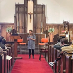 Amy Julia peaking at the front of the church sanctuary, which has traditional architecture, featuring wooden pews, high ceilings with exposed beams, and large hanging lanterns.