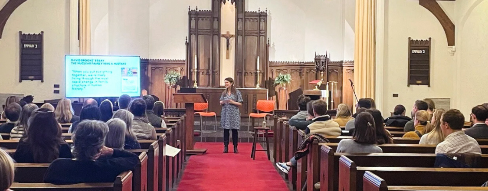 Amy Julia peaking at the front of the church sanctuary, which has traditional architecture, featuring wooden pews, high ceilings with exposed beams, and large hanging lanterns.