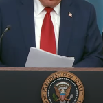 A cropped image of President Trump standing behind a podium with the seal of the President of the United States. He is wearing a dark suit, a white shirt, and a red tie, holding a piece of paper. The background features the American flag and the presidential flag.