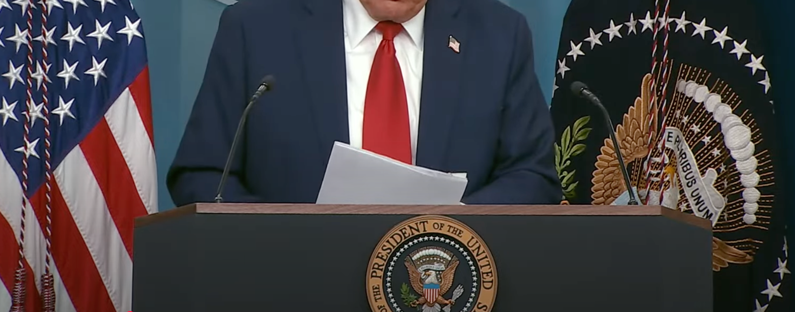 A cropped image of President Trump standing behind a podium with the seal of the President of the United States. He is wearing a dark suit, a white shirt, and a red tie, holding a piece of paper. The background features the American flag and the presidential flag.