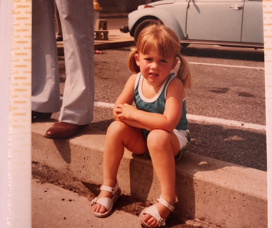 Amy Julia as a young girl with her blonde hair in pigtails. She’s sitting on a concrete curb in a parking lot, slightly squinting at the camera with a serious expression. A vintage light blue vehicle is parked in the background.