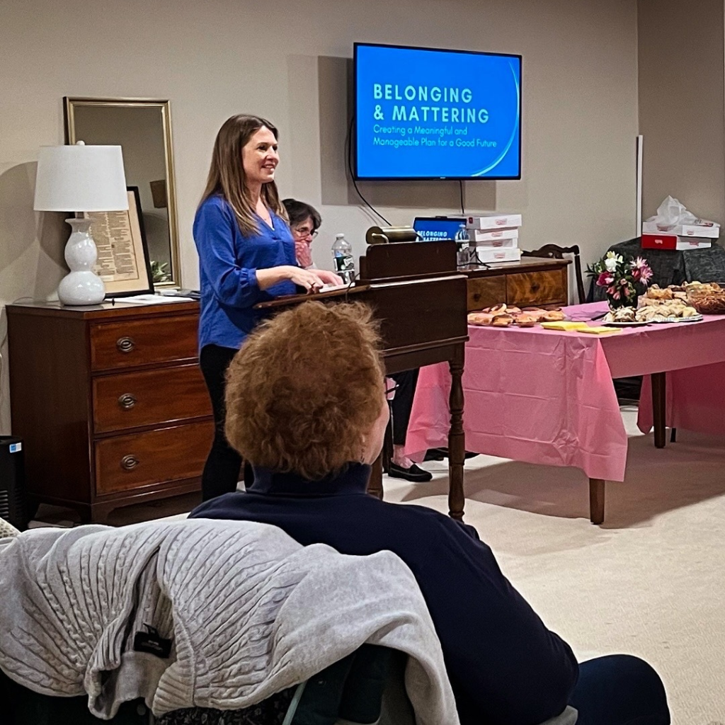 I stand at a wooden podium, speaking to an audience in a cozy room. A screen behind me displays the presentation title: "Belonging & Mattering: Creating a Meaningful and Manageable Plan for a Good Future."