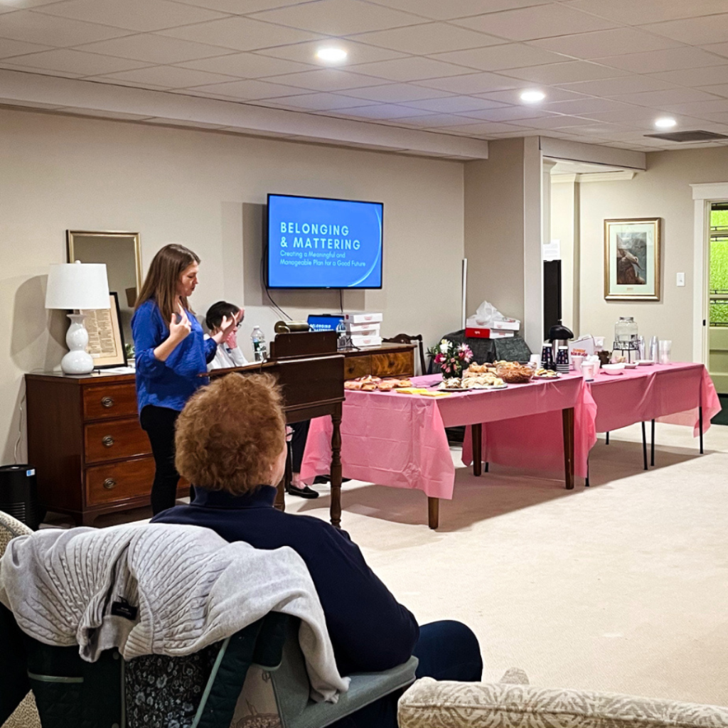 I stand at a wooden podium, speaking to an audience in a cozy room. A screen behind me displays the presentation title: "Belonging & Mattering: Creating a Meaningful and Manageable Plan for a Good Future."