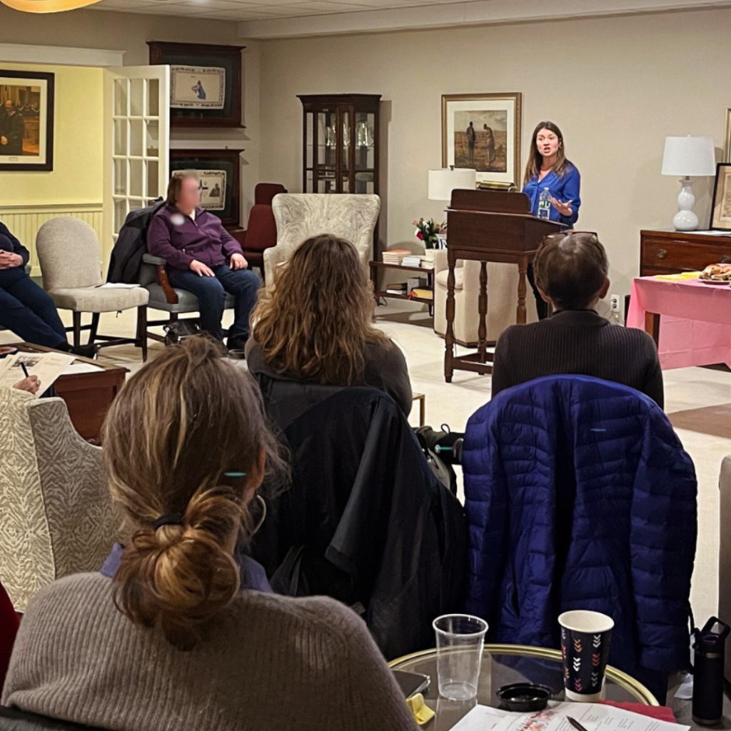 Amy Julia, stands at a wooden podium, speaking to a seated audience in a warmly lit room.