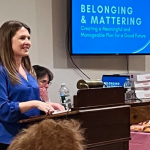 I stand at a wooden podium, speaking to an audience in a cozy room. A screen behind me displays the presentation title: "Belonging & Mattering: Creating a Meaningful and Manageable Plan for a Good Future."