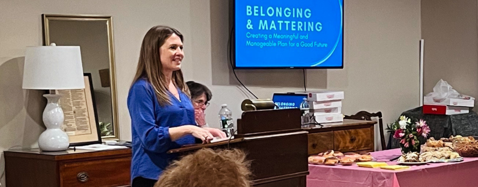 I stand at a wooden podium, speaking to an audience in a cozy room. A screen behind me displays the presentation title: "Belonging & Mattering: Creating a Meaningful and Manageable Plan for a Good Future."