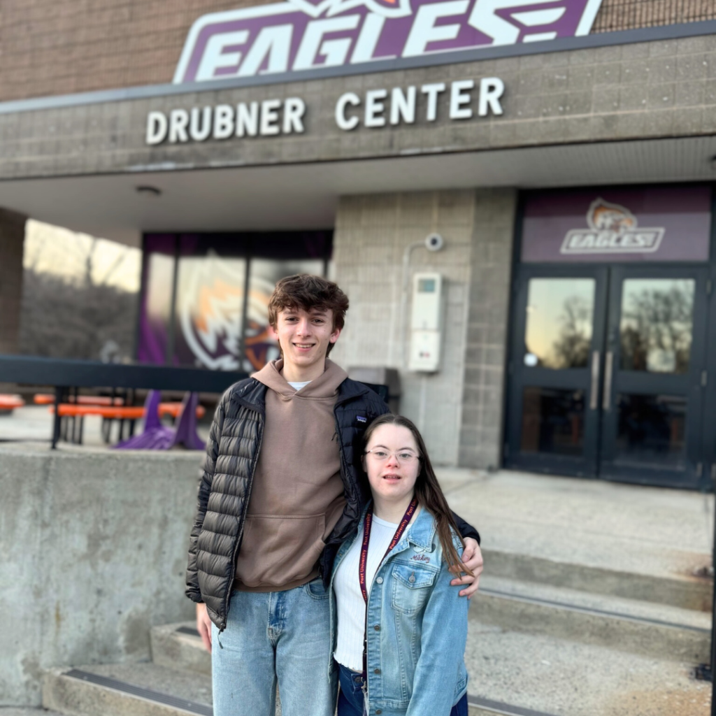 William and Penny stand together outside the Drubner Center at Post University. They are smiling, and William has his arm around Penny’s shoulders